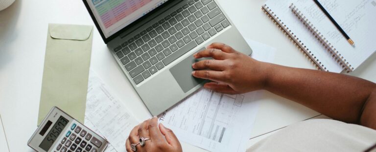 Pessoa organizando seus investimentos em uma mesa. Ela está utilizando uma calculadora, notebook e papéis.
