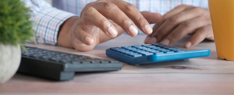Pessoa usando a calculadora em uma mesa. Ao lado há um notebook e uma parte de uma xícara amarela.