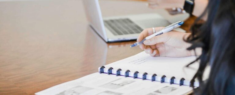 Pessoa escrevendo em um caderno em uma mesa. Um notebook também está na mesa.