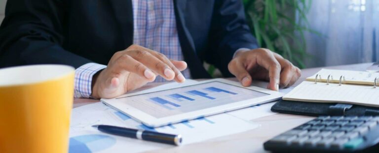 Homem analisando um gráfico em um tablet que está em uma mesa. Na mesa, também estão canetas, uma xícara amarela, uma calculadora e uma agenda.